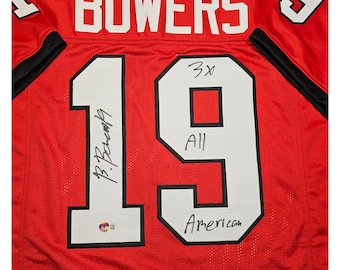May include: Red football jersey with the name "BOWERS" and the number "19" on the back. The jersey is signed by the player with the text "B. Bowers" and "All American".