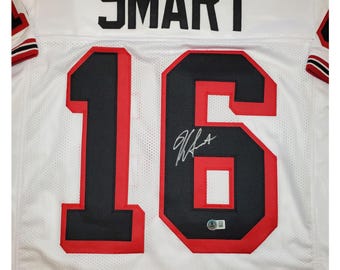 May include: White football jersey with black and red trim. The number 16 is on the back of the jersey with a black outline and red trim. The name "SMART" is above the number. The jersey is signed in black ink.