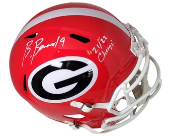 May include: A red and white football helmet with a black and white 'G' logo. The helmet is signed 'B.  Bann 19' and '21/22 Champs'.