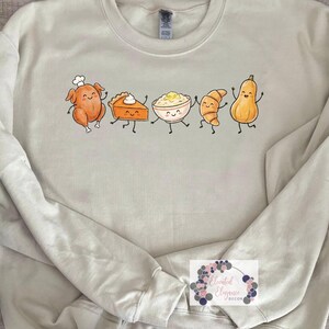 May include: A beige crewneck sweatshirt featuring a graphic print of five cartoon food items with smiling faces: a roasted turkey wearing a chef hat, a slice of pumpkin pie, a bowl of mashed potatoes with butter, a croissant, and a butternut squash.