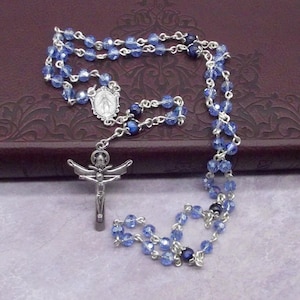 May include: A rosary featuring blue crystal beads and silver-coloured metal chain links. It includes a crucifix and a religious emblem. The faceted beads reflect light. This is a religious item.