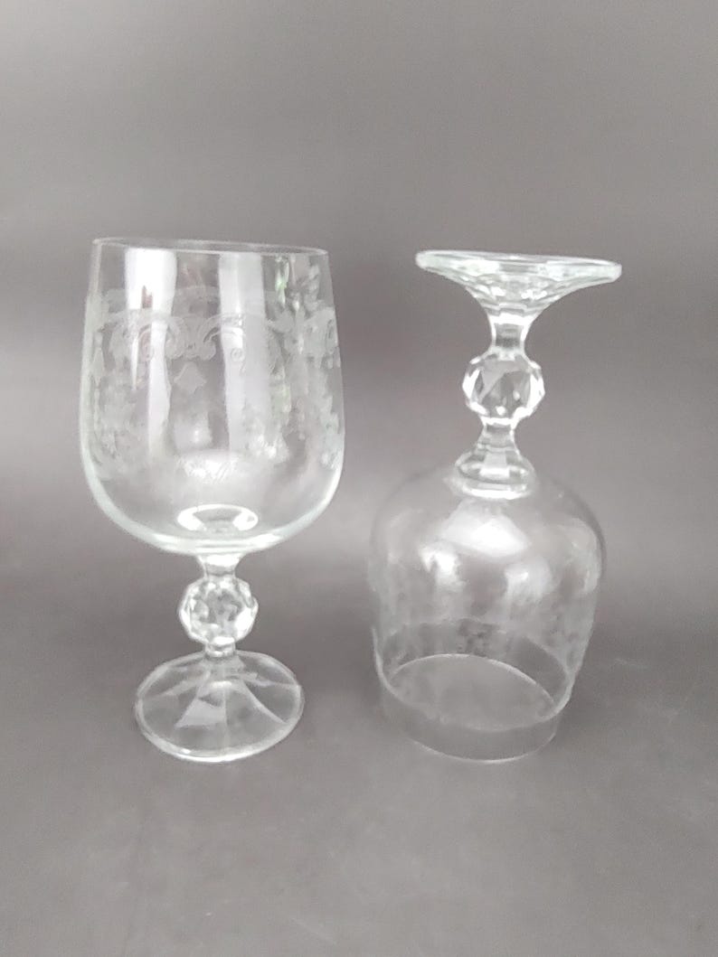 May include: Two clear, stemmed wine glasses. One glass is upright, the other is upside down. The upright glass has etched floral designs on the bowl. Both glasses have a faceted ball on the stem and a round base.