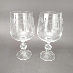 May include: Two clear wine glasses with etched floral designs. Each glass has a faceted crystal-like stem and a round base. The glasses are displayed on a gray background.