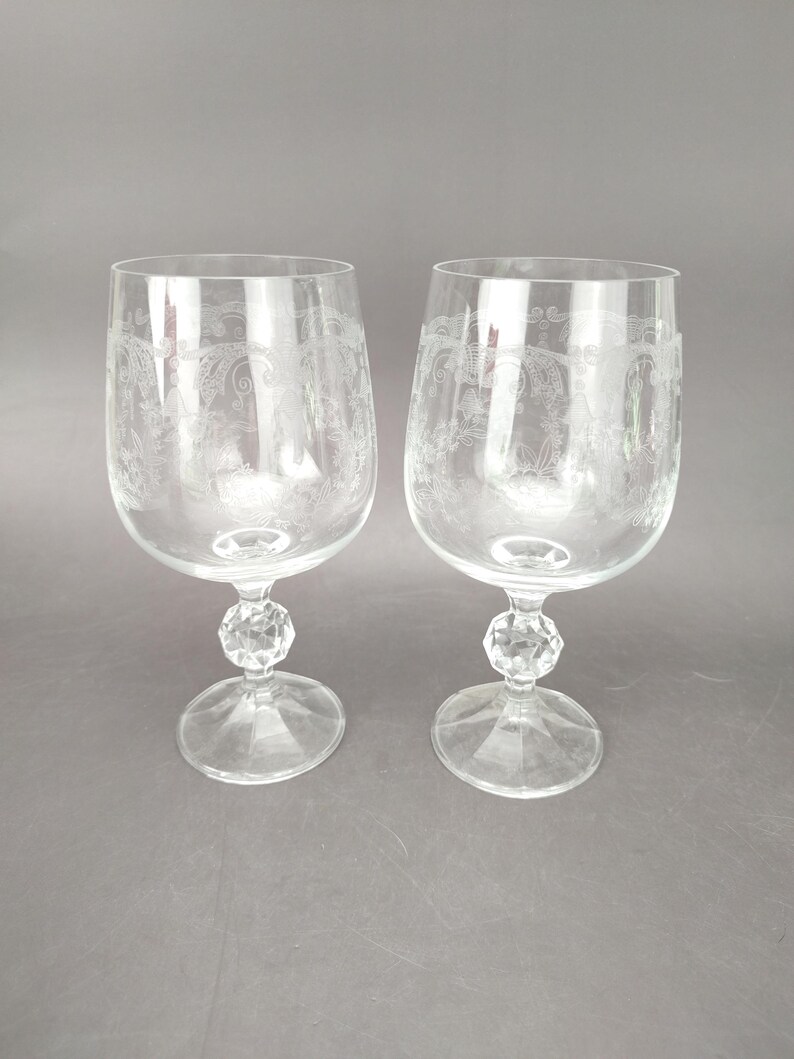 May include: Two clear wine glasses with etched floral designs. Each glass has a faceted crystal-like stem and a round base. The glasses are displayed on a gray background.