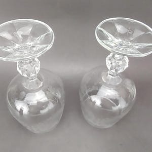 May include: Two clear, stemmed wine glasses with a unique design. The bowls are teardrop-shaped, and the stems feature a faceted crystal-like detail. The rims are wide and circular, with a starburst pattern emanating from the center.
