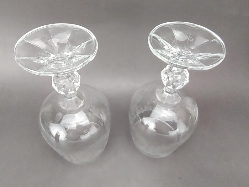 May include: Two clear, stemmed wine glasses with a unique design. The bowls are teardrop-shaped, and the stems feature a faceted crystal-like detail. The rims are wide and circular, with a starburst pattern emanating from the center.