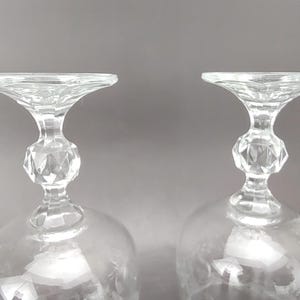 May include: Two clear crystal wine glasses. The glasses have a rounded bowl, a faceted stem, and a flared rim. The stem has a diamond-shaped design. The glasses are set against a gray background.