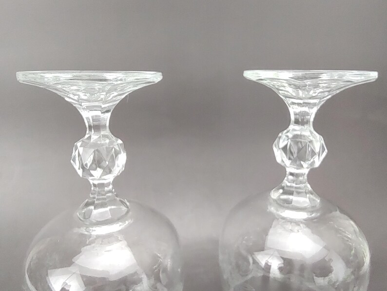 May include: Two clear crystal wine glasses. The glasses have a rounded bowl, a faceted stem, and a flared rim. The stem has a diamond-shaped design. The glasses are set against a gray background.