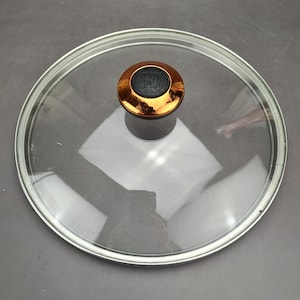 May include: A clear glass pot lid with a shiny silver rim. The lid features a copper-colored handle with the letters "FD" embossed on a black background. The lid is round and appears to be new.