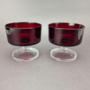 May include: Two stemmed dessert glasses. The bowls are a deep red color, and the stems and bases are clear. The glasses have a simple, elegant design, perfect for serving desserts or small portions.