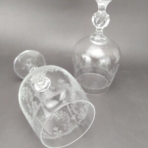 May include: Two clear, stemmed wine glasses with etched floral designs. One glass is upright, the other is lying on its side. The stems have a faceted detail and a flared top. The glasses are on a gray background.