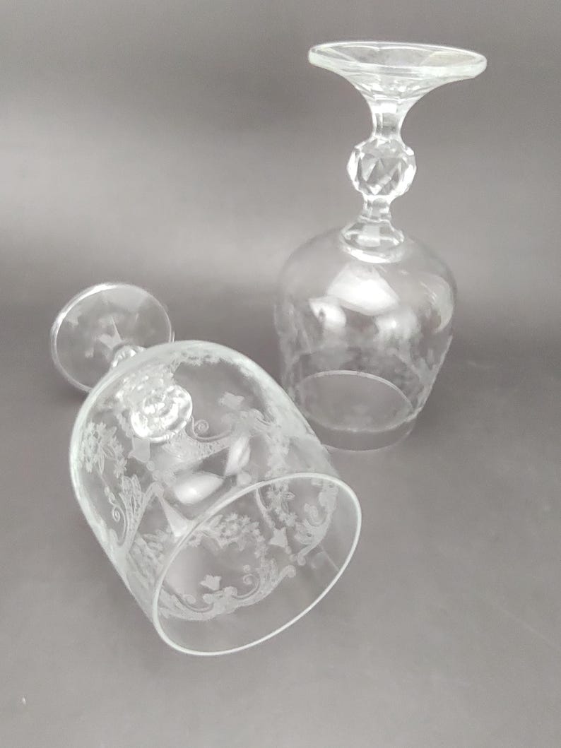 May include: Two clear, stemmed wine glasses with etched floral designs. One glass is upright, the other is lying on its side. The stems have a faceted detail and a flared top. The glasses are on a gray background.