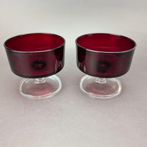May include: Two matching dessert bowls made of deep red glass. Each bowl features a wide, rounded basin supported by a short, thick stem that connects to a clear, circular glass base, providing stability for the dark red glass vessels.