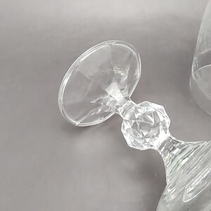 May include: Clear glass stemware with a faceted stem and a wide, shallow bowl. The stemware is lying on its side, showcasing its design. A second glass with etched floral details is partially visible.