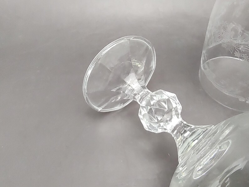 May include: Clear glass stemware with a faceted stem and a wide, shallow bowl. The stemware is lying on its side, showcasing its design. A second glass with etched floral details is partially visible.