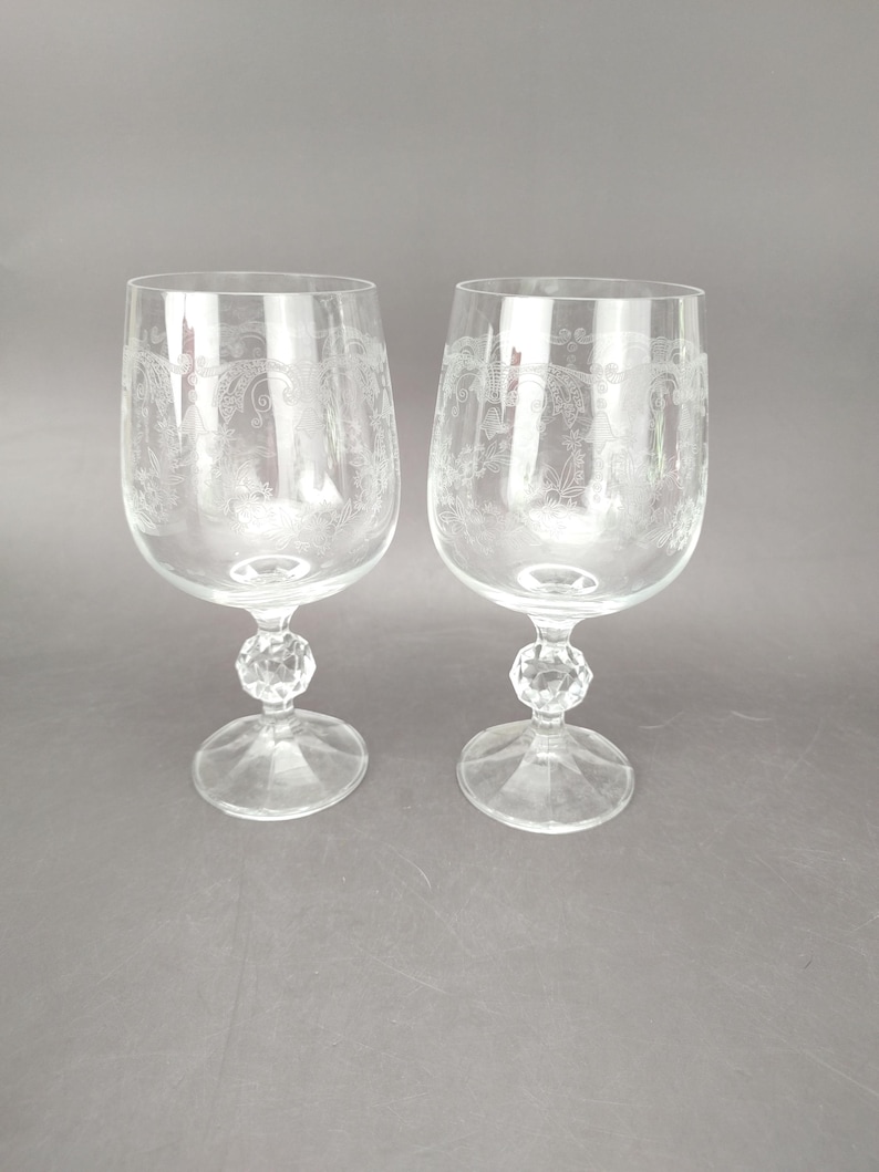 May include: Two clear wine glasses with etched floral designs. The glasses have a rounded bowl, a faceted stem, and a circular base. The background is a solid gray.
