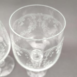 May include: Two clear, stemmed glasses with etched floral and scrollwork designs. The glasses have a wide bowl and a rounded base. The etched details are visible around the rim and the lower portion of the bowl.