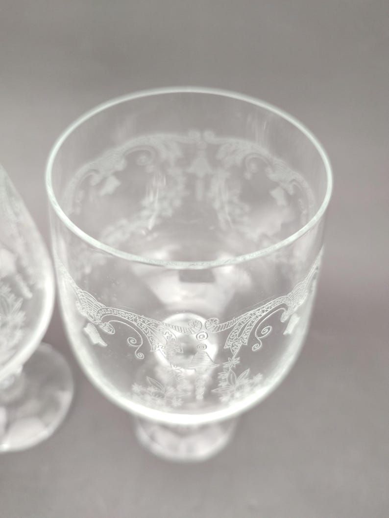 May include: Two clear, stemmed glasses with etched floral and scrollwork designs. The glasses have a wide bowl and a rounded base. The etched details are visible around the rim and the lower portion of the bowl.