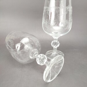 May include: Two clear, stemmed wine glasses with etched floral designs. One glass is upright, the other is lying on its side. The bases and stems have a faceted, geometric design. The glasses are on a gray surface.