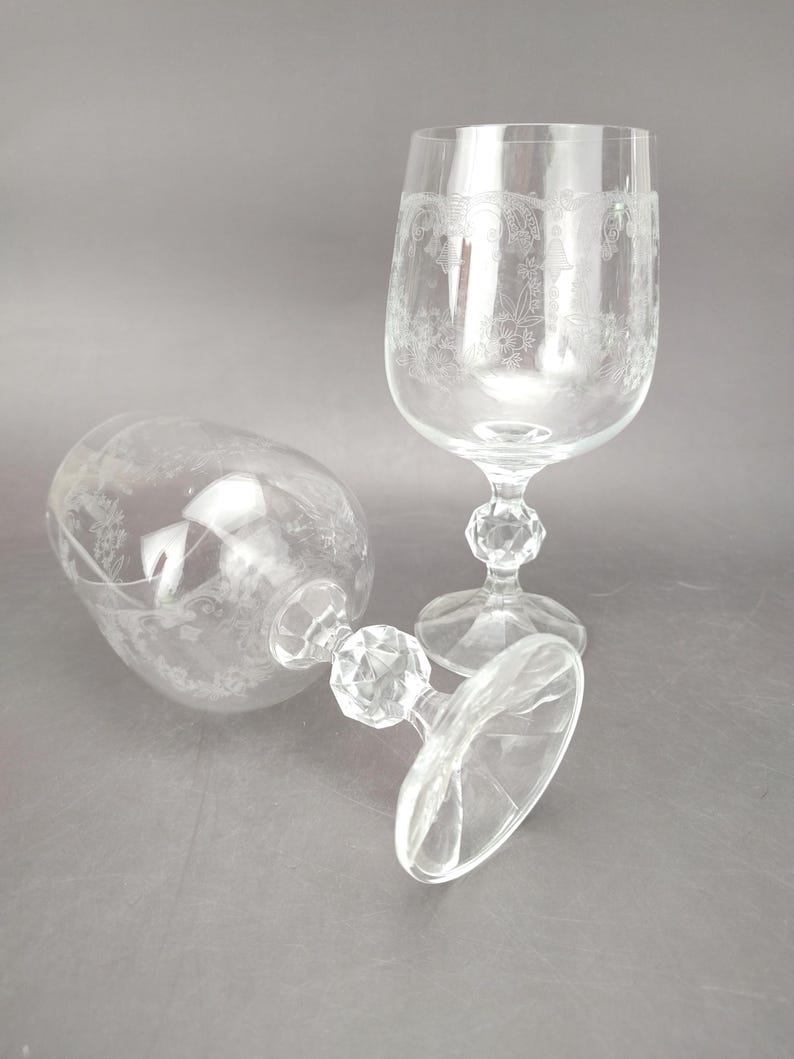 May include: Two clear, stemmed wine glasses with etched floral designs. One glass is upright, the other is lying on its side. The bases and stems have a faceted, geometric design. The glasses are on a gray surface.