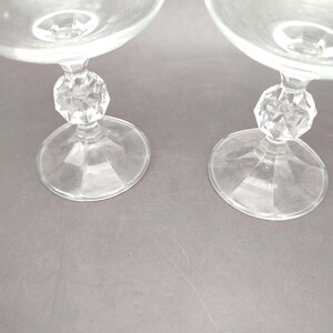 May include: Two clear glass goblets with a round bowl and a faceted, crystal-like stem. The goblets have a wide, circular base. The stem has a decorative, geometric design. The glasses are transparent and appear to be made of glass.