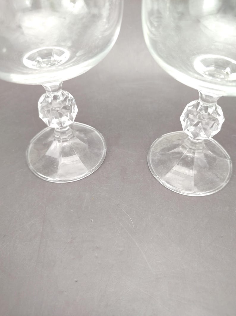 May include: Two clear glass goblets with a round bowl and a faceted, crystal-like stem. The goblets have a wide, circular base. The stem has a decorative, geometric design. The glasses are transparent and appear to be made of glass.