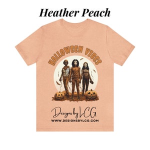 May include: A heather peach t-shirt with a graphic of three figures in front of a moon with the text "Halloween Vibes" in a circle around the moon. The text "Designs by LCG" and "www.designsbylcg.com" are below the graphic.