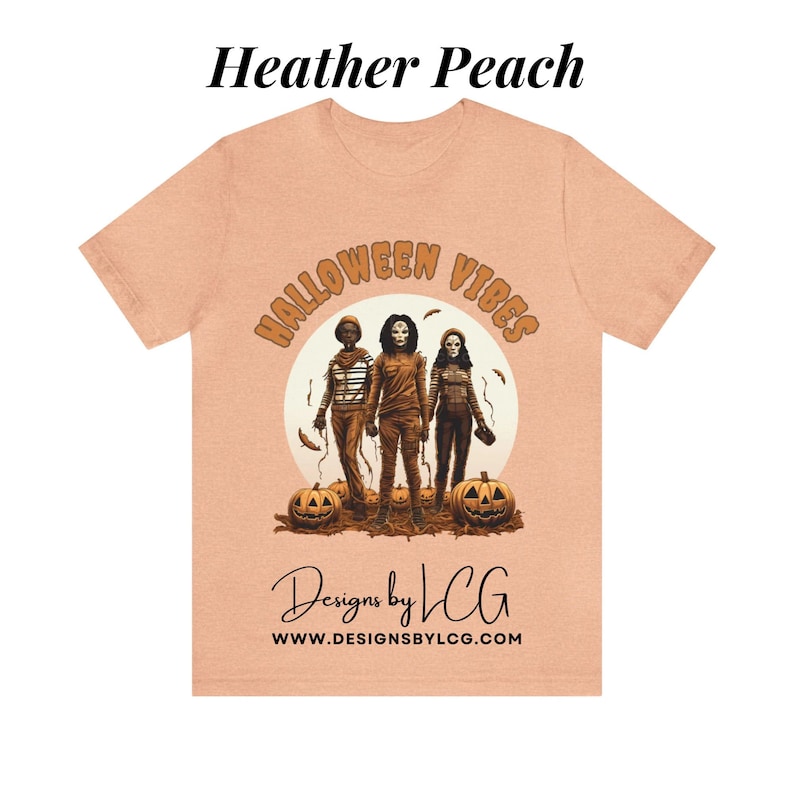 May include: A heather peach t-shirt with a graphic of three figures in front of a moon with the text "Halloween Vibes" in a circle around the moon. The text "Designs by LCG" and "www.designsbylcg.com" are below the graphic.