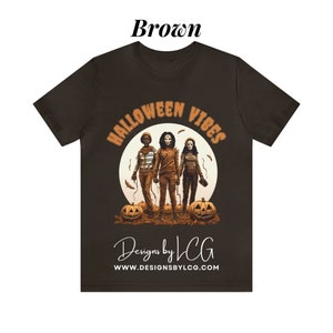 May include: Brown t-shirt with a graphic of three people in front of a moon with the text "Halloween Vibes" and "Designs by LCG" with the website address "www.designsbylcg.com".