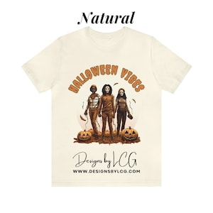 May include: A white t-shirt with a graphic print of three people in Halloween costumes. The text "Halloween Vibes" is in orange and brown letters above the graphic. The text "Designs by LCG" and "www.designsbylcg.com" is below the graphic.
