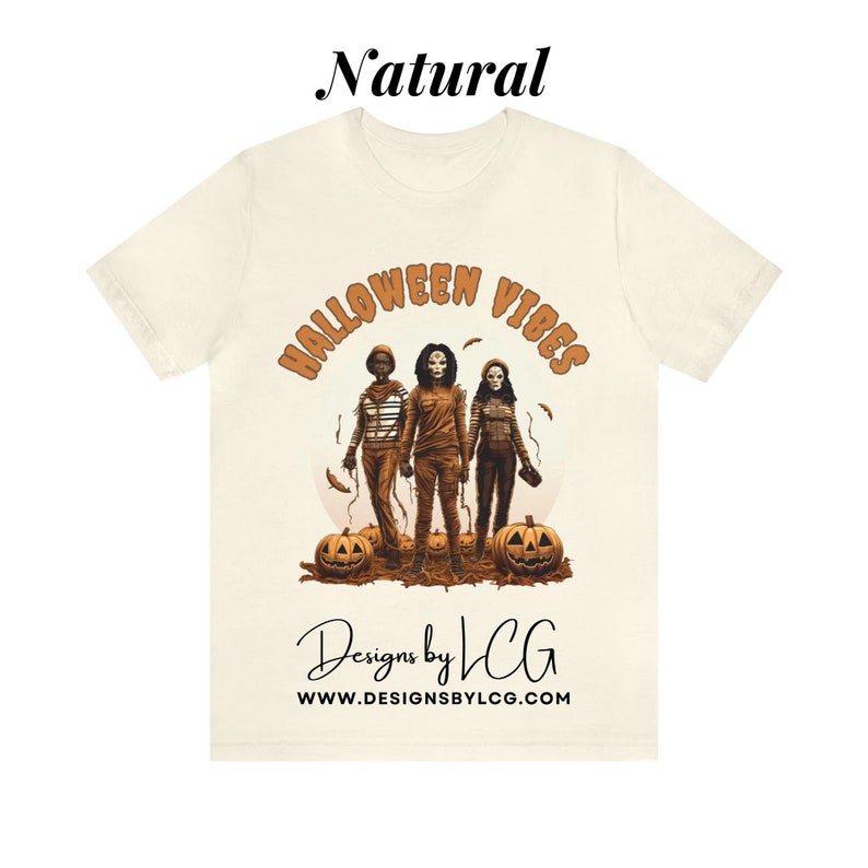 May include: A white t-shirt with a graphic print of three people in Halloween costumes. The text "Halloween Vibes" is in orange and brown letters above the graphic. The text "Designs by LCG" and "www.designsbylcg.com" is below the graphic.