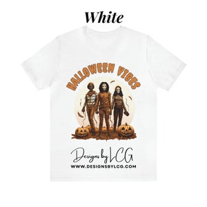 May include: White t-shirt with a graphic of three people in Halloween costumes, pumpkins, and the text "Halloween Vibes" and "Designs by LCG www.designsbylcg.com".