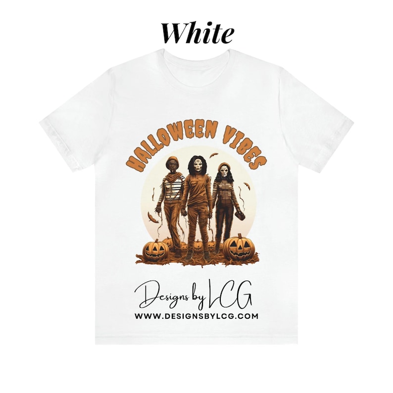 May include: White t-shirt with a graphic of three people in Halloween costumes, pumpkins, and the text "Halloween Vibes" and "Designs by LCG www.designsbylcg.com".