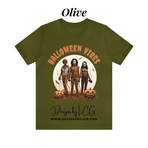May include: Olive green t-shirt with a graphic of three figures in front of a full moon with the text "Halloween Vibes" in orange and black. The text "Designs by LCG" and "www.designsbylcg.com" are printed below the graphic.