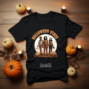 May include: Black t-shirt with a graphic of three figures in front of a full moon. The text "Halloween Vibes" is in an orange arch above the figures. The text "Designs by JCG" and a website address are at the bottom of the shirt.