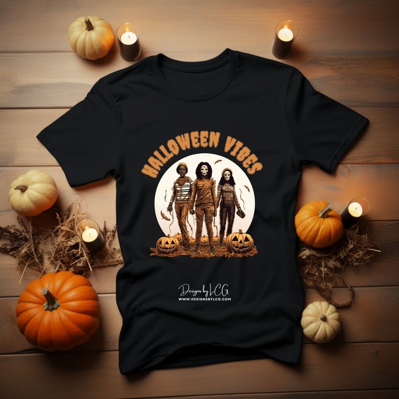 May include: Black t-shirt with a graphic of three figures in front of a full moon. The text "Halloween Vibes" is in an orange arch above the figures. The text "Designs by JCG" and a website address are at the bottom of the shirt.