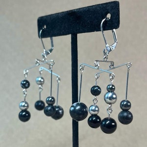 May include: A pair of silver earrings with black beads hanging from a mobile-like structure. The earrings are on a black display stand.