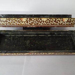 May include: A rectangular wooden box with a black interior and a decorative floral pattern on the top and bottom edges. The pattern is in shades of brown, gold, and white.