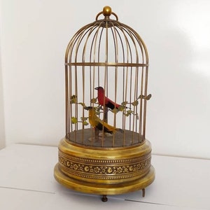 May include: A vintage brass birdcage with two mechanical birds inside. The birdcage has a round base with an ornate design and a domed top. The birds are red and yellow and are perched on a branch.