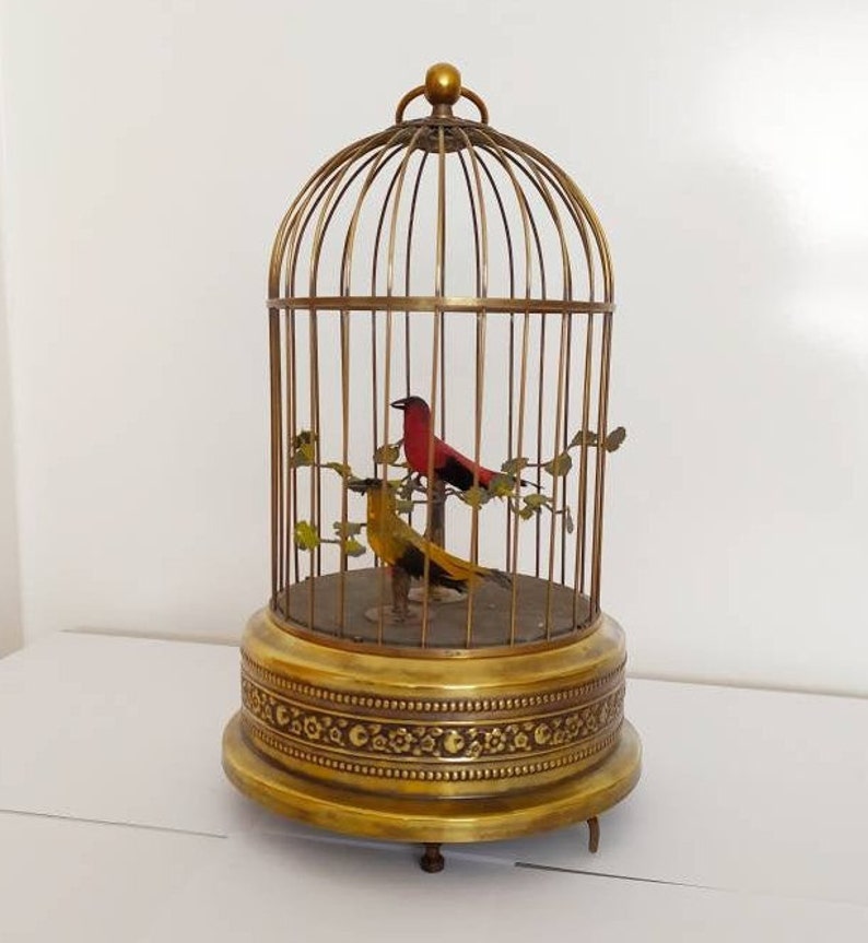 May include: A vintage brass birdcage with two mechanical birds inside. The birdcage has a round base with an ornate design and a domed top. The birds are red and yellow and are perched on a branch.