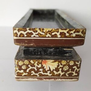 May include: A two-tiered wooden box with intricate floral and geometric patterns painted in gold, white, and red on a brown background. The box has a rectangular shape and a raised lid.