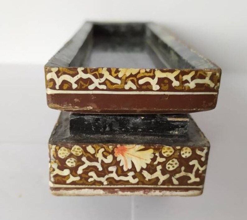 May include: A two-tiered wooden box with intricate floral and geometric patterns painted in gold, white, and red on a brown background. The box has a rectangular shape and a raised lid.