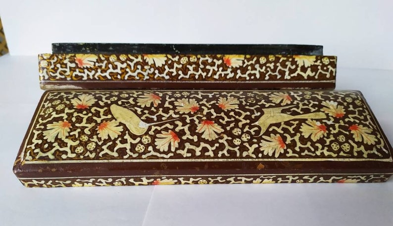 May include: A brown wooden box with a decorative floral design in gold and red. The box has a hinged lid and is decorated with intricate patterns.