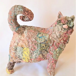 May include: A colourful ceramic cat sculpture with a textured finish. The cat is predominantly pink, with accents of blue, yellow, and brown. Its tail is curled. The sculpture has a handmade look, with visible brushstrokes and a playful design.