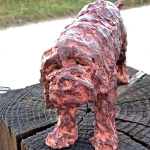 May include: A sculpture of a dog with a textured, reddish-brown surface. The figure is depicted in a standing position, showcasing a rough, hand-modeled appearance with deep grooves and ridges across its entire body, including the head and legs.