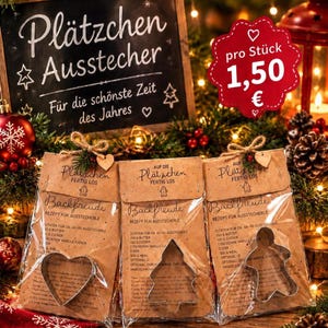 Könnte beinhalten: Drei braune Papierverpackungen mit Metall-Ausstechformen in Herz-, Tannenbaum- und Lebkuchenmann-Form. Jede Packung ist mit dem Text 'Backfreude' und einem Rezept bedruckt, mit Kordel verschnürt und mit einem kleinen Holzherz verziert.
