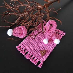 Könnte beinhalten: Ein rosa gehäkeltes Set aus Schal, Mütze und Fäustlingen mit weißen Bommeln. Das Set ist mit einem kleinen Holzhäuschen verziert.