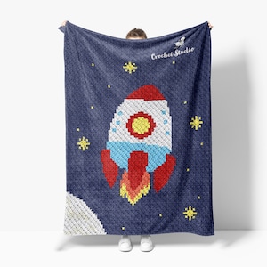 May include: A navy blue crocheted blanket featuring a pixelated rocket ship design with red, white, blue, and yellow accents. The blanket also includes yellow stars and a crescent moon against a dark blue background. The text "Crochet Studio" is visible.