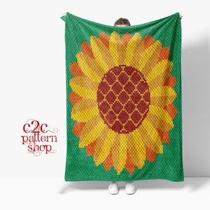 Op de afbeelding: Een gehaakte deken met een groot zonnebloemmotief. De zonnebloem is gemaakt van verschillende tinten geel, oranje en rood. Het midden van de zonnebloem is donkerrood met een geometrisch patroon. De deken is felgroen.