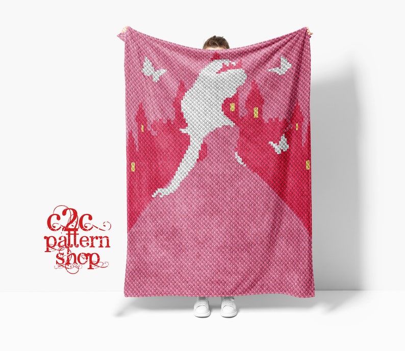 May include: A pink crochet blanket with a white silhouette of a castle and a flying figure. The blanket is held up by a person, and the text "C2C pattern shop" is visible in the lower left corner.